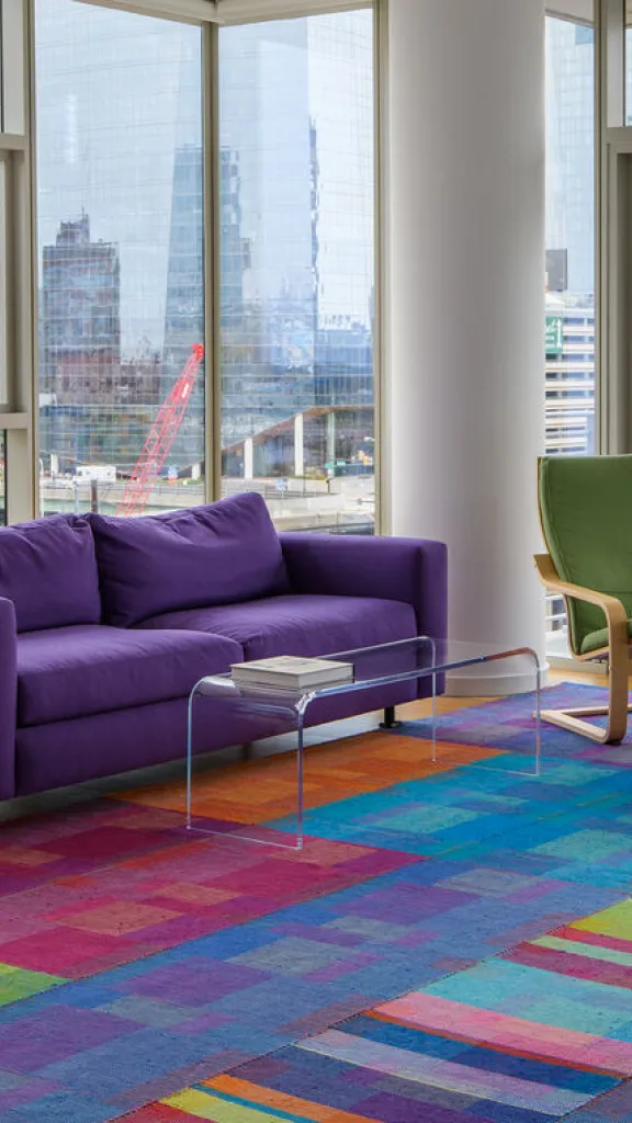 A handwoven rug that is green, red, purple, and blue in a room with a purple couch and green chair, with windows all around overlooking a city skyline