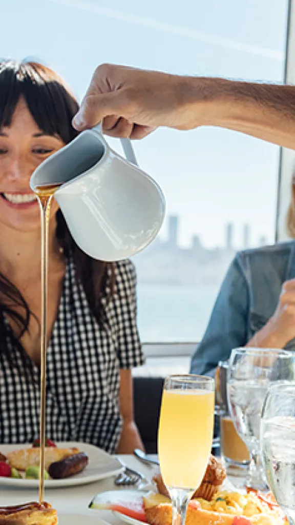 People sitting at a table eating brunch with mimosas. They are watching someone pour syrup on their food. 