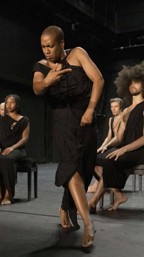 Four performers sitting heads held high observing the dancer in front in the midst of gesturing and gliding to their left. 