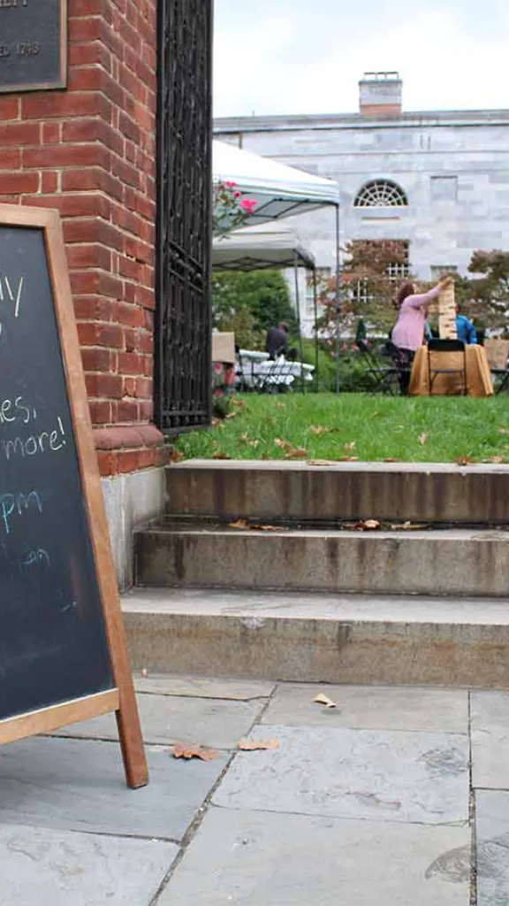 Sidewalk with chalkboard sandwich board.  Boad says "APS Family Fun Day Activities, Games, Snacks, and More! 12pm - 4pm" with a drawing of an apple. People in the garden in the background playing games. 