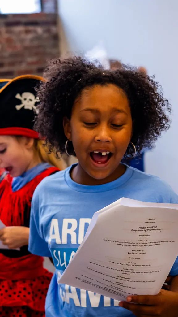 Two children read scripts. Girl in front is wearing a blue shirt. The boy in the back is dressed as a pirate.