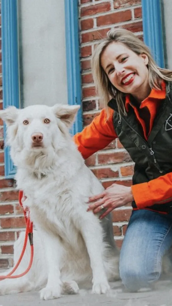 smiling dog trainer kneeling down with cute white dog, pops of orange and blue against a brick building as the backdrop