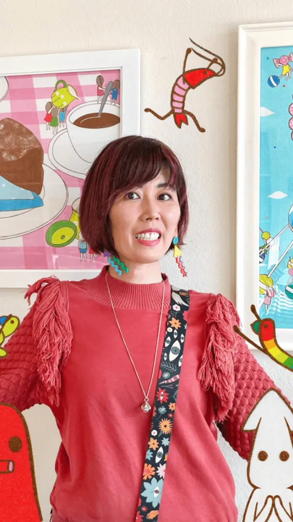 Photo of artist Naoshi with two framed pieces of art hanging on a wall behind her, cartoon characters surround her face 