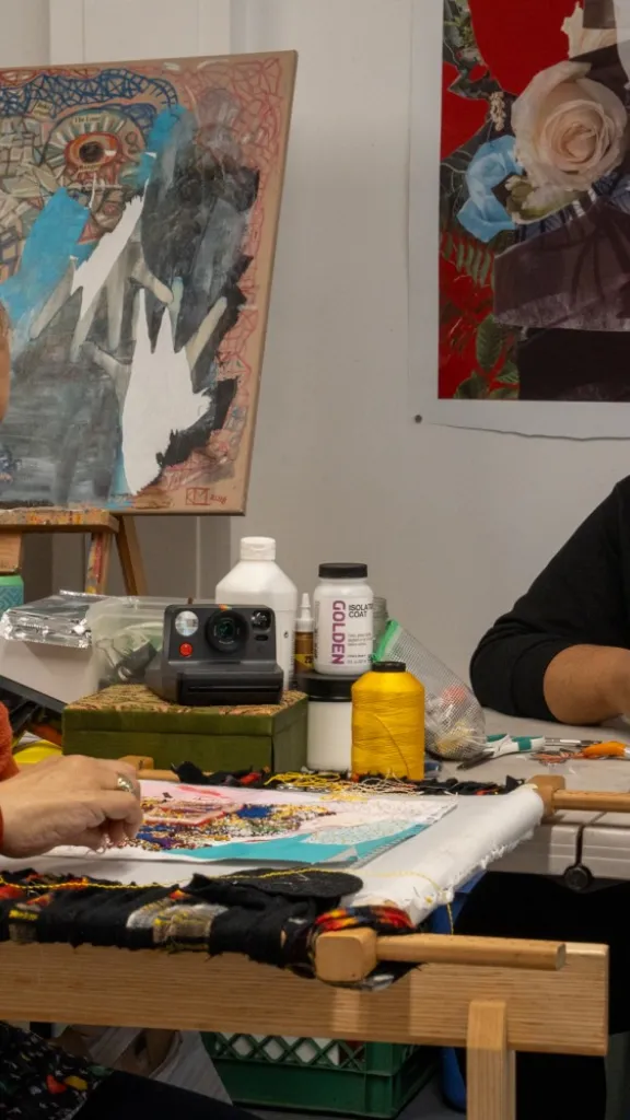 GrioXArts working on a mixed-media piece in her studio, surrounded by paintings and art supplies.