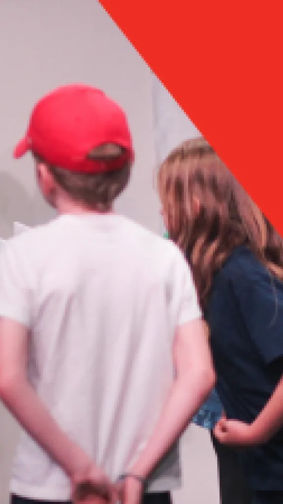 A boy in a green striped shirt holds a paper in his hand and makes a funny, scrunched face. Other children hold papers in a similar way. Text that reads "Play-in-a-day" and "Grades K-5."