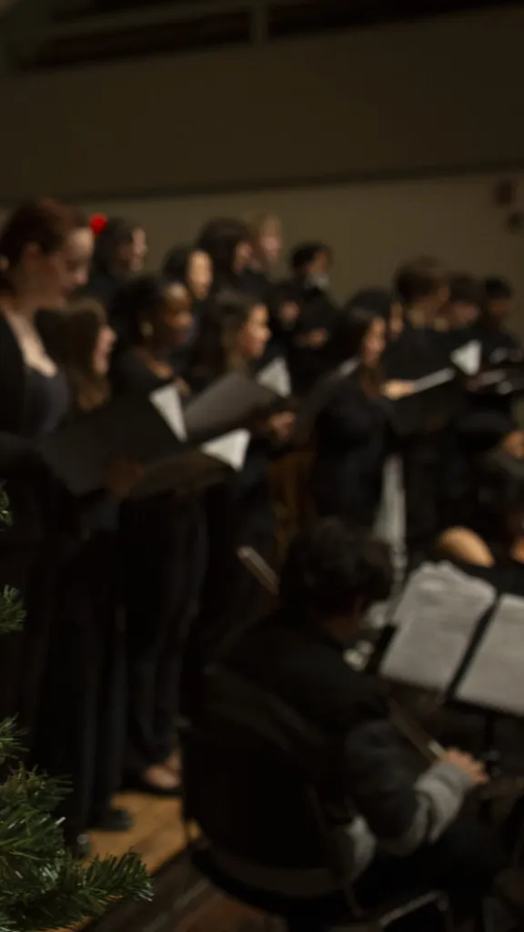 A Christmas tree with lights and ornaments is shown in the foreground. In the background, a choir and orchestra perform.