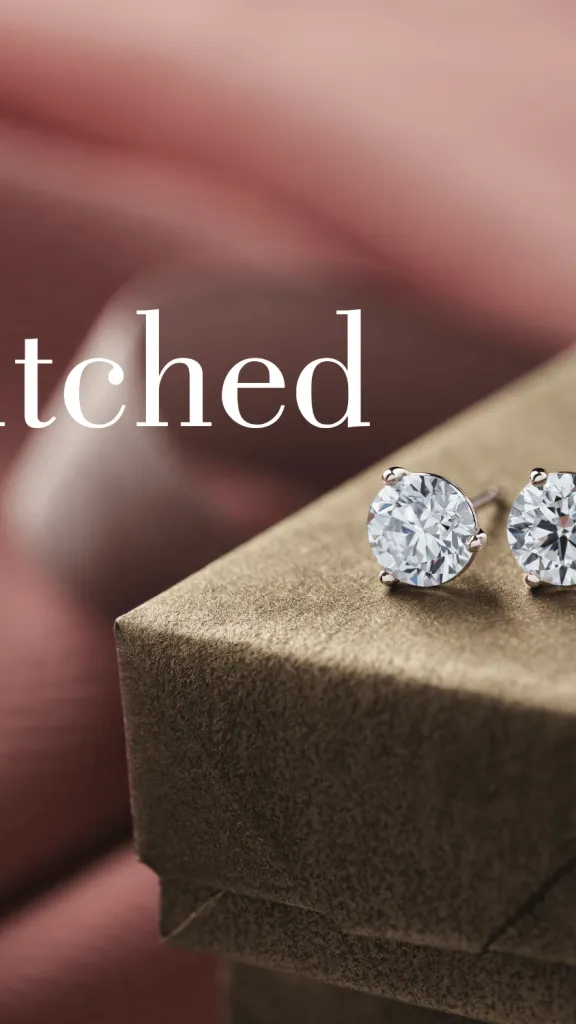 A pair of diamond studs sitting on a green gift box and surrounded by pink fabric and ribbon