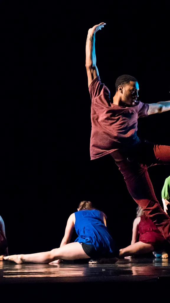 Contemporary dancers freeze in dynamic motion on a dark stage, one leaping midair as others flow low to the floor.