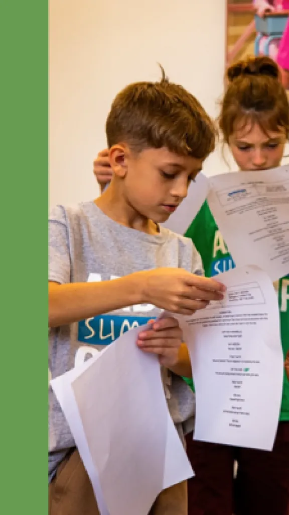 Two children holding scripts next to a kneeling teacher that is assisting them. Next to the image is the text: 'Arden Summer Camp, Play Practice, One Week Specialty Camp for Grades 1-5