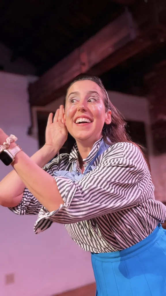 Lee Minora smiling, she gestures animatedly with one hand cupped to her ear and the other extended outward.