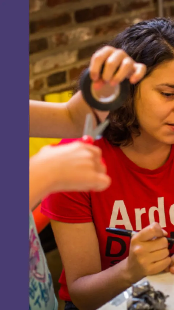 A Woman is holding a pen and sitting with two young students; they are all holding art materials and crafting. Next to the image, the text reads: 'Arden Summer Camp, Stage Makeup, One Day Masterclass for Grades 9-12.' 