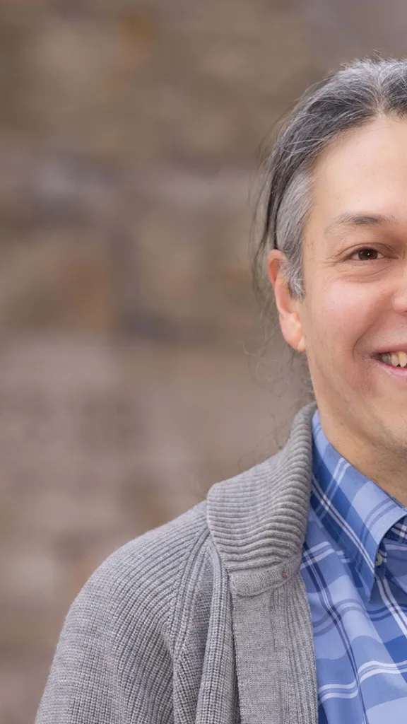 Graphic of book to the left of man with grey hair, blue shirt, and grey sweater