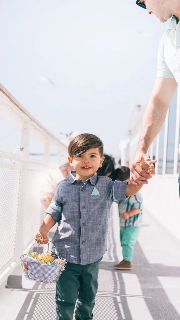 A small child holding a woven basket walks hand‑in‑hand with an adult along a sunlit outdoor deck.