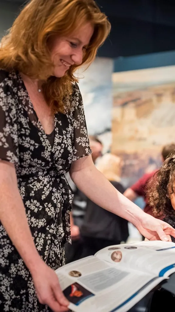 Woman with red hair in brown and white dress holds paper for young girl with curly brown hair to stamp
