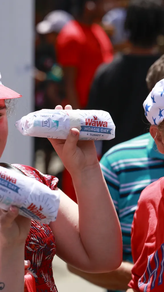 Three individuals wearing Wawa hats and holding wrapped hoagies