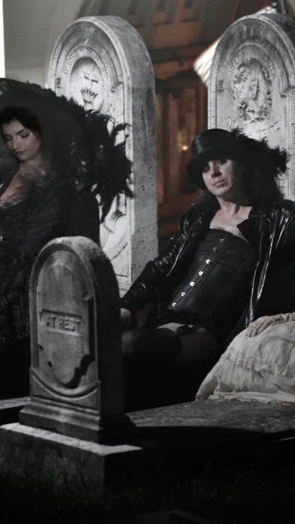 One man and two women, all dressed in Gothic costumes, lie on three gravestones in a graveyard.