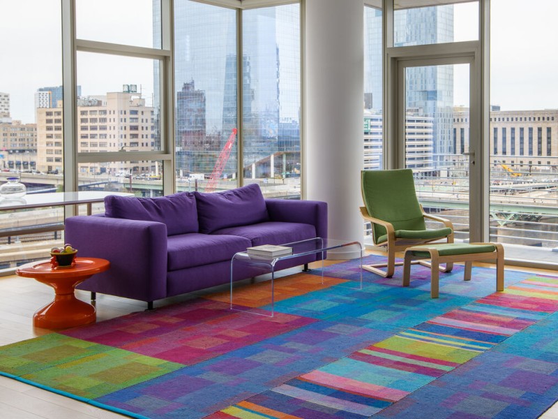 A handwoven rug that is green, red, purple, and blue in a room with a purple couch and green chair, with windows all around overlooking a city skyline