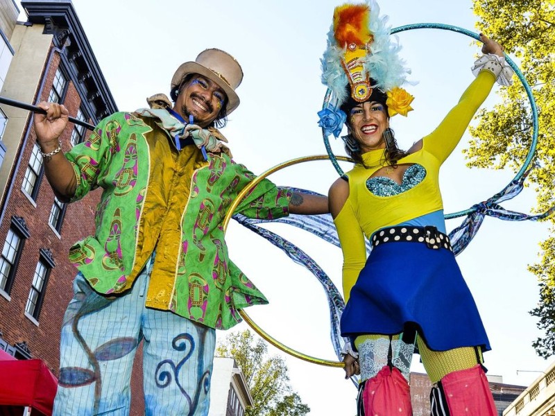 Two stilt walkers in bright, festive costumes holding hula hoops and smiling at Old City Fest