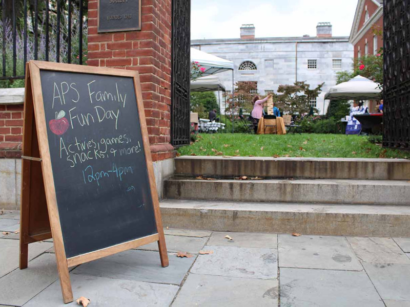 Sidewalk with chalkboard sandwich board.  Boad says "APS Family Fun Day Activities, Games, Snacks, and More! 12pm - 4pm" with a drawing of an apple. People in the garden in the background playing games. 
