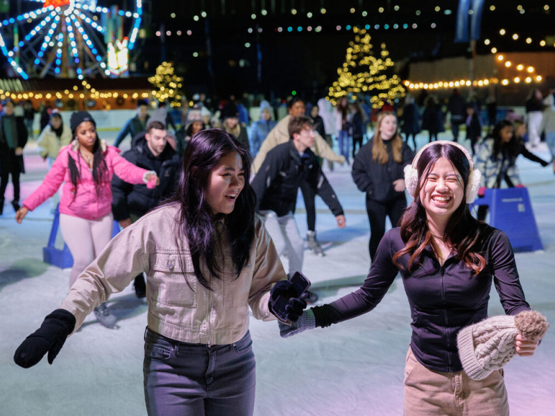 Photo of people skating, laughing and having fun. A ferris wheel is pictured in the background.