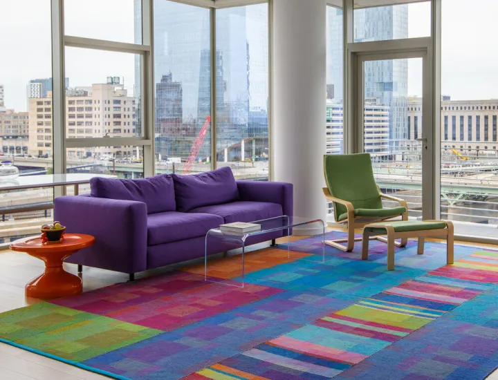 A handwoven rug that is green, red, purple, and blue in a room with a purple couch and green chair, with windows all around overlooking a city skyline
