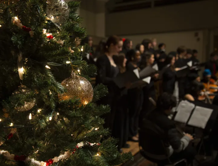 A Christmas tree with lights and ornaments is shown in the foreground. In the background, a choir and orchestra perform.
