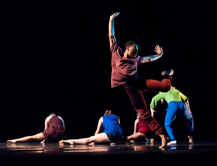 Contemporary dancers freeze in dynamic motion on a dark stage, one leaping midair as others flow low to the floor.