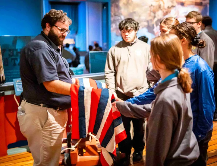 Tour guide in navy shirt and khaki pants shows teenagers a replica US flag
