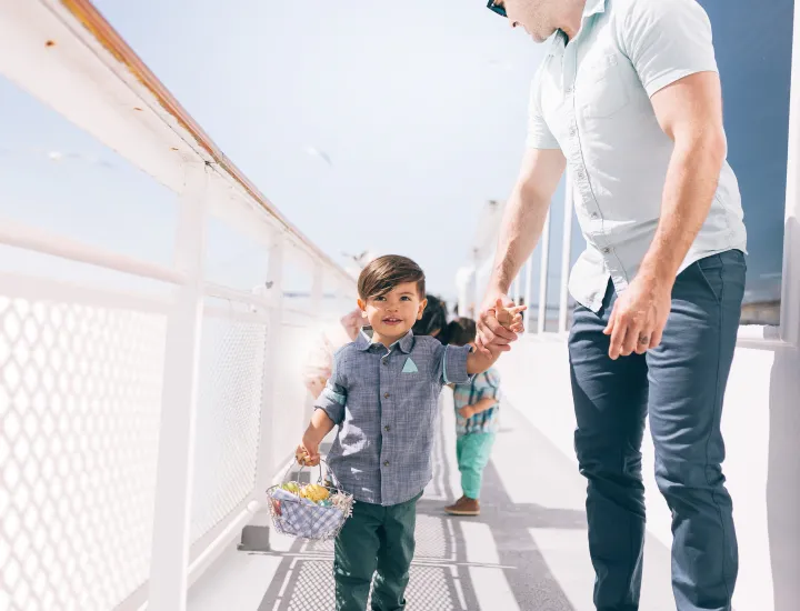 A small child holding a woven basket walks hand‑in‑hand with an adult along a sunlit outdoor deck.