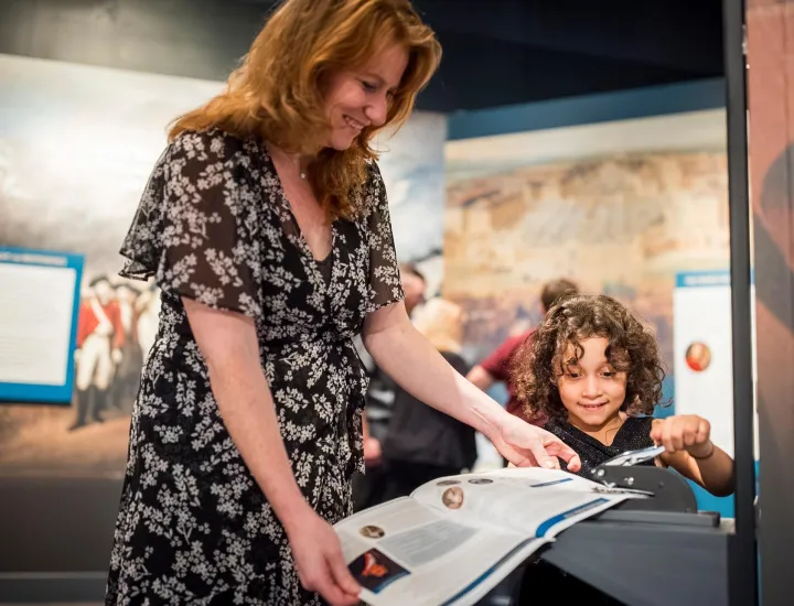 Woman with red hair in brown and white dress holds paper for young girl with curly brown hair to stamp