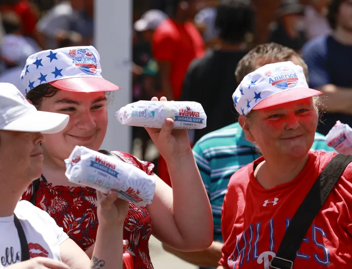 Three individuals wearing Wawa hats and holding wrapped hoagies