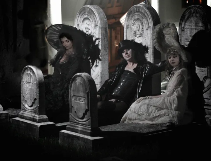 One man and two women, all dressed in Gothic costumes, lie on three gravestones in a graveyard.