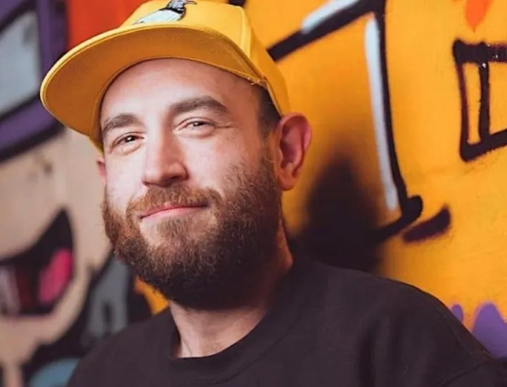 Photo of Eitan Levine, a white man with a brown beard wearing a yellow baseball cap and a black shirt standing in front of a graffitied wall